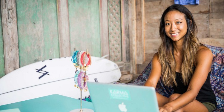 Image of Cordia sitting in a tropical room on her laptop with a surfboard leaning against the wall in the background